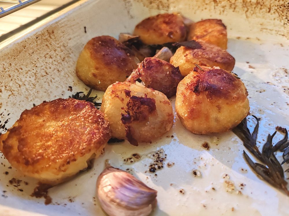 8 golden and crispy roast potatoes with rosemary and garlic in a white enamel dish 