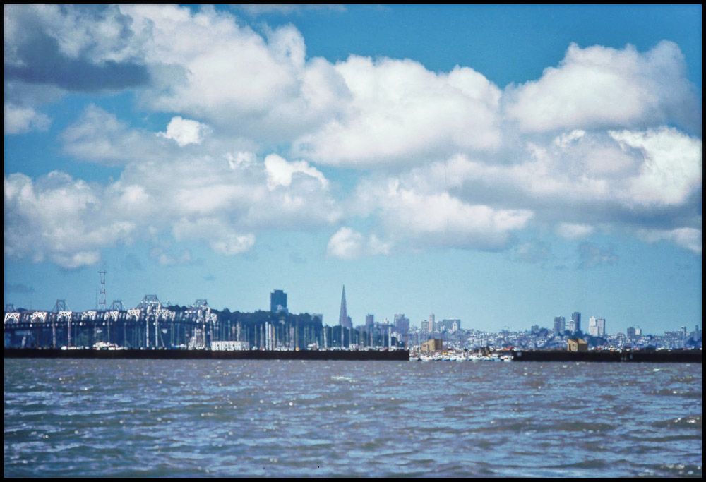 ~1970 Looking out across the San Francisco Bay for a glimpse of the San Francisco skyline back in the day. Blue sky with fluffy white clouds. 
Haven't been back in ages, but I'm wondering it that was the end of the Bay Bridge that had problems during the last (?) earthquake?