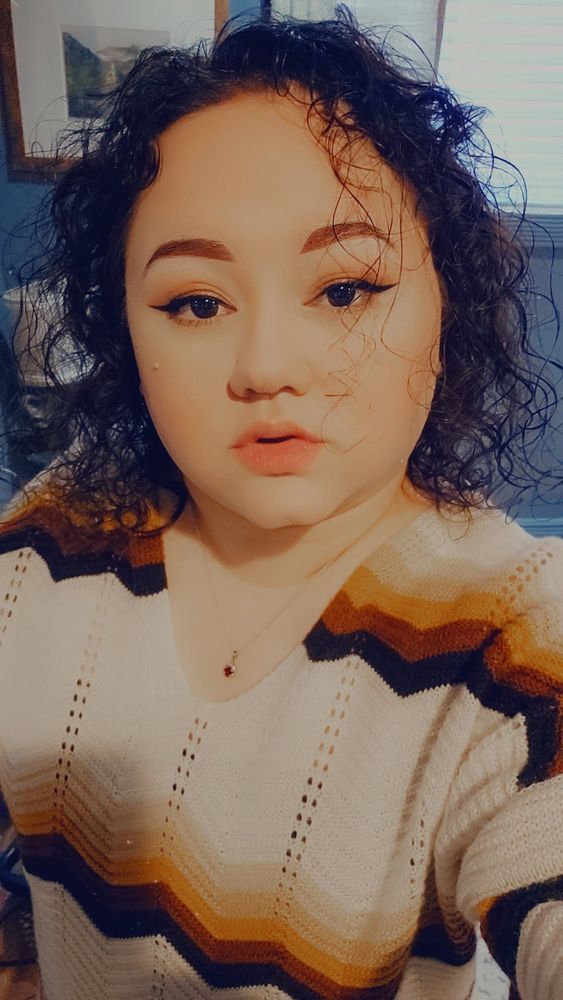 A selfie of user Neon Gravestone. Their hair is almost black & they are wearing a white sweater with beige, orange, and black zigzag. Their makeup is done 