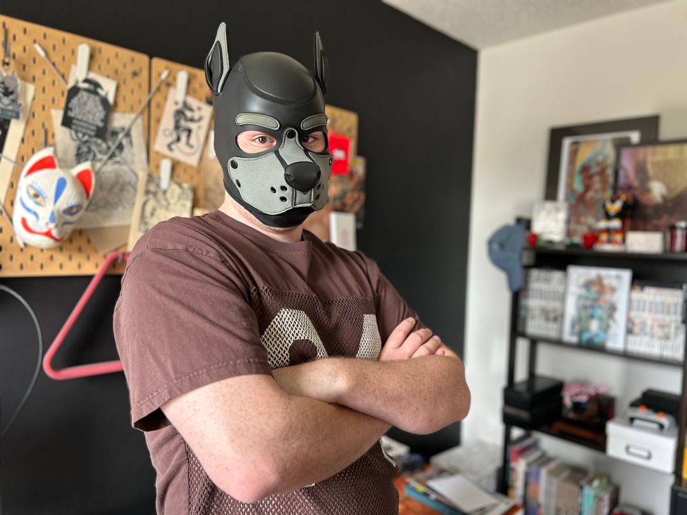 a man in a dog mask standing with arms crossed in front of books, papers, and poster arts