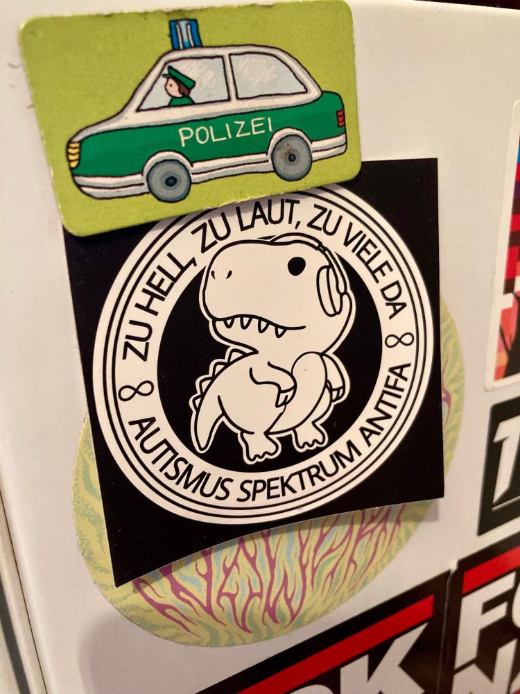 Ein Antifa-Logo mit einem Dinosaurier mit Gehörschutz und der Aufschrift "Zu hell, zu laut, zu viele da - Autismus Spektrum Antifa"