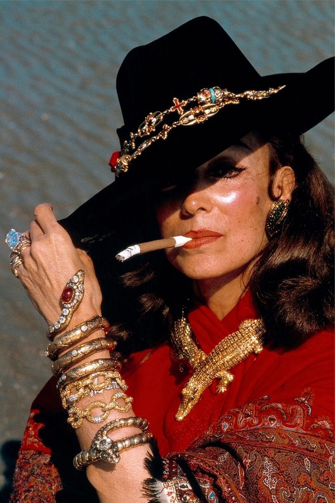 Maria Felix wears a broad-brimmed black hat cocked over one eye; it has jewelry around the crown. Her hair is down and she wears a red top and a beautiful red paisley shawl. She has a half-smoked cigarillo in a holder clutched in her teeth. 
