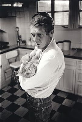 Steve McQueen in his kitchen holding a marmalade (? b&w) cat in his arms like a baby. The cat's head is visible over McQueen's arm and it looks peaceful. McQueen, in a white shirt and jeans, looks pensive. 