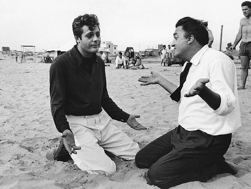 Marcello (dark shirt, white pants) is kneeling on the beach, with his hands held palms up in a classically Italian gesture of resignation. A cigarette is going in his right hand. Fellini, in a white shirt, dark tie and dark pants, is also kneeling and appears to be showing his star how to do the palms-up shrug. Only Fellini's gesture is even more "what can you do?" (b&w)