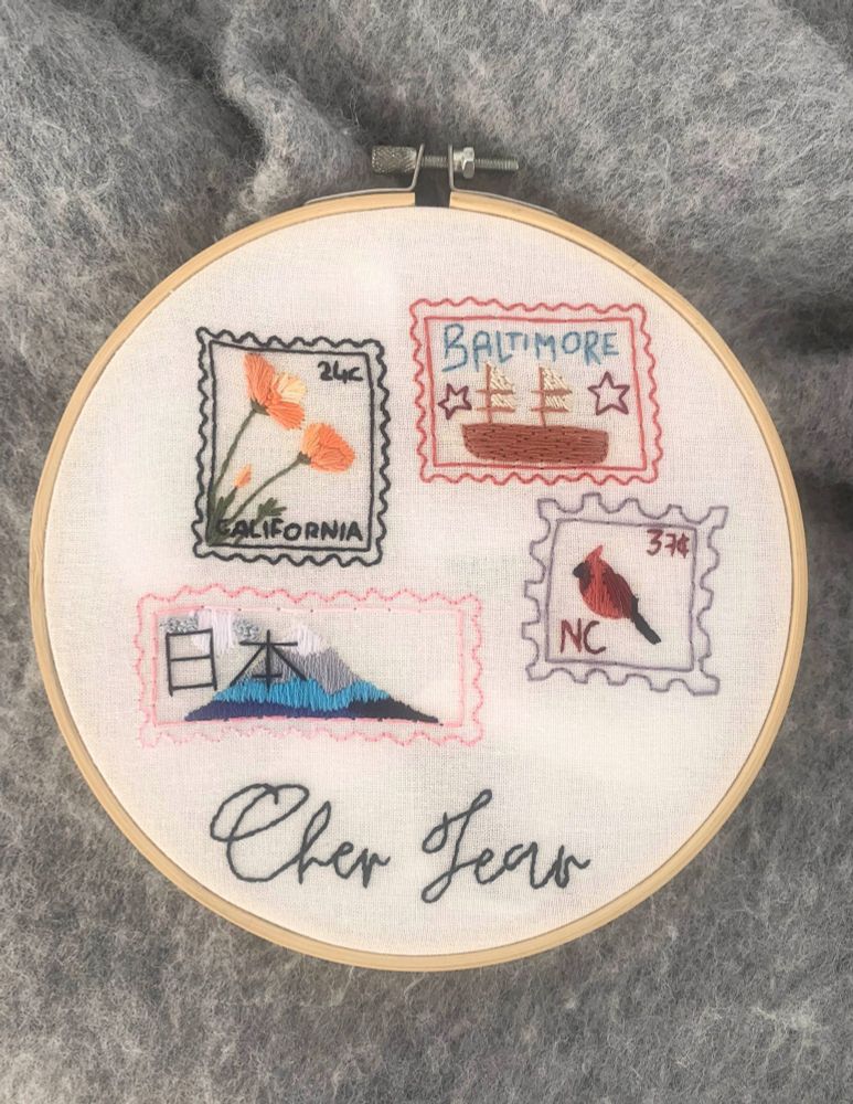 photograph of an embroidery piece showing four different stamps (california with california poppies on the top left, baltimore with a ship and two stars on top right, japan with mount fuji on bottom left, and north carolina with a red cardinal on bottom right.) and the words Cher Jean across the bottom of the piece in a calligraphy style font