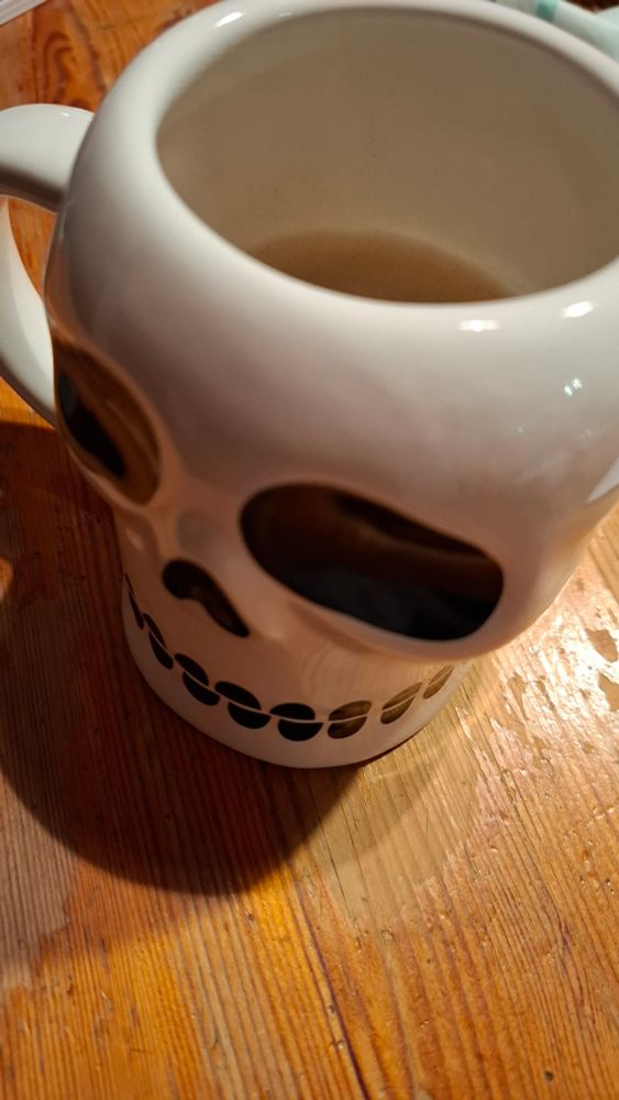 Eine weiße Kaffeetasse in angedeuteter Schädelform mit aufgemalten Augen- und Nasenhöhlen und Zähnen in schwarz auf einem alten Holztisch 
