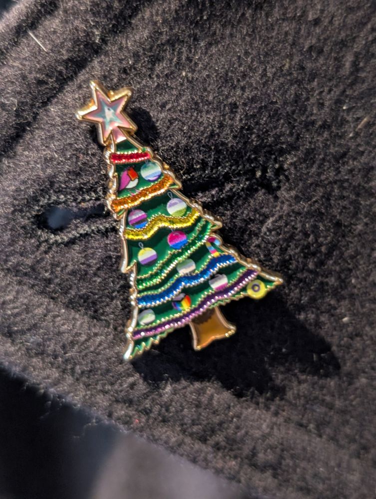A gold enamel pin of a green Christmas tree covered in multi-coloured tinsel and featuring LGBTQ+ themed baubles and a trans flag star