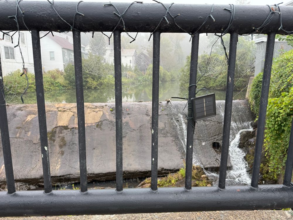 A black fence is covered in lights that are turned off. There’s a river behind the fence, with fog over the river. VERY faint spiderwebs are in between the bars of the fence.