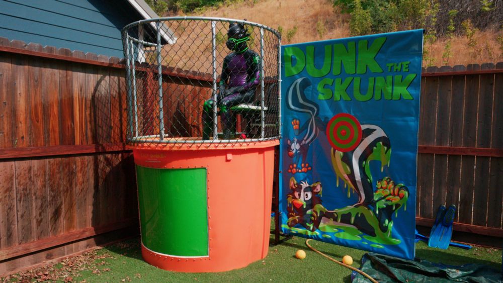 a rubber dragon sits at the top of a dunk tank full of green slime. the blue banner to his left says "dunk the skunk" in green text with a picture of a skunk covered in green slime on it.