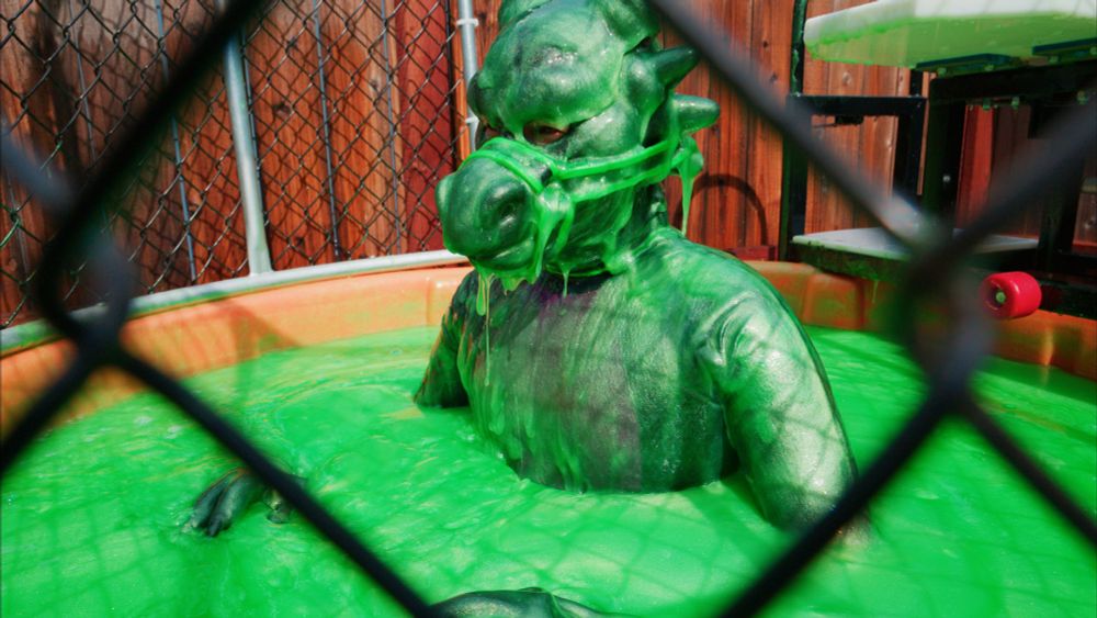 a rubber dragon standing in a tank of bright green slime as slime drips off of his snout and down his chest