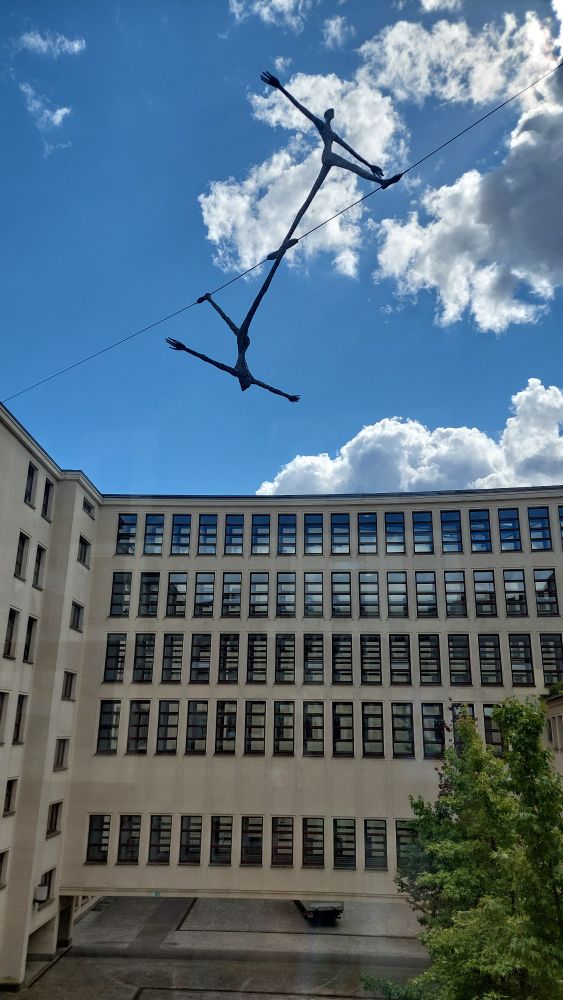 Kunstobjekt unter freiem Himmel. Ein Mensch balanciert mit ausgestreckten Armen auf einem Seil in luftiger Höhe, darunter ist kopfüber der gleiche Mensch. Im Hintergrund sind Gebäude.