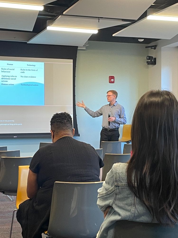 Vertical image of a book talk. In the foreground, there is an audience member with long black hair and another audience member with short hair and a face mask. Jeffrey Boase is talking in the background and gesturing toward a PowerPoint presentation.