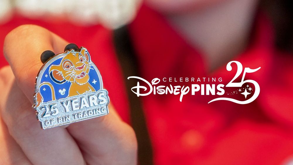 A close-up of a hand holding a Disney pin featuring young Simba from The Lion King on a blue background with stars and Mickey icons. The bottom of the pin reads "25 YEARS OF PIN TRADING" in silver text. The hand is in front of a blurred red background, possibly a Disney Cast Member uniform. To the right, the text "Celebrating Disney Pins 25" appears in a stylized white font with sparkles, commemorating the 25th anniversary of Disney's pin trading tradition.