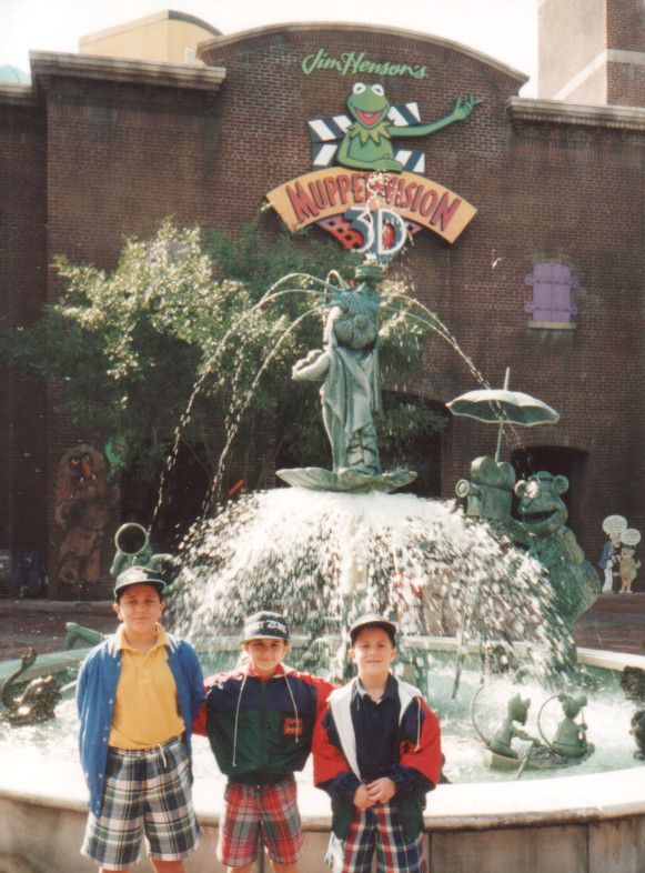 A nostalgic photo of three children standing in front of the iconic fountain outside the Jim Henson’s MuppetVision 3D* attraction at Disney’s Hollywood Studios. The fountain features Miss Piggy dressed as the Statue of Liberty, surrounded by other Muppet characters in humorous poses, with water spraying from various parts of the sculpture. The children are wearing colorful 1990s outfits, including plaid shorts and windbreakers, along with baseball caps. Behind them, the red-brick building displays the attraction’s sign featuring Kermit the Frog, with the name “Jim Henson’s Muppet*Vision 3D” prominently displayed. Trees and whimsical decorations add to the lively atmosphere of the scene.