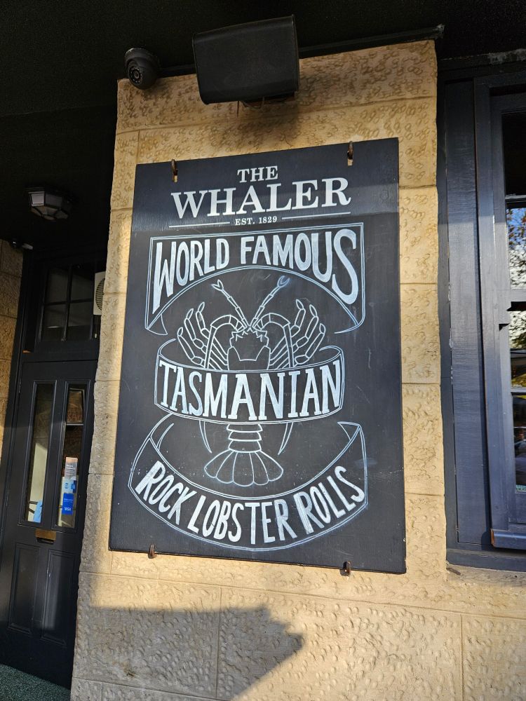A chalkboard style sign on a pale sandstone wall outside a pub. The heading says "The Whaler - est. 1829" and underneath is a white line-art drawing of a lobster, around and under which are white line-art drawn banners which read "world famous Tasmanian rock lobster rolls". 