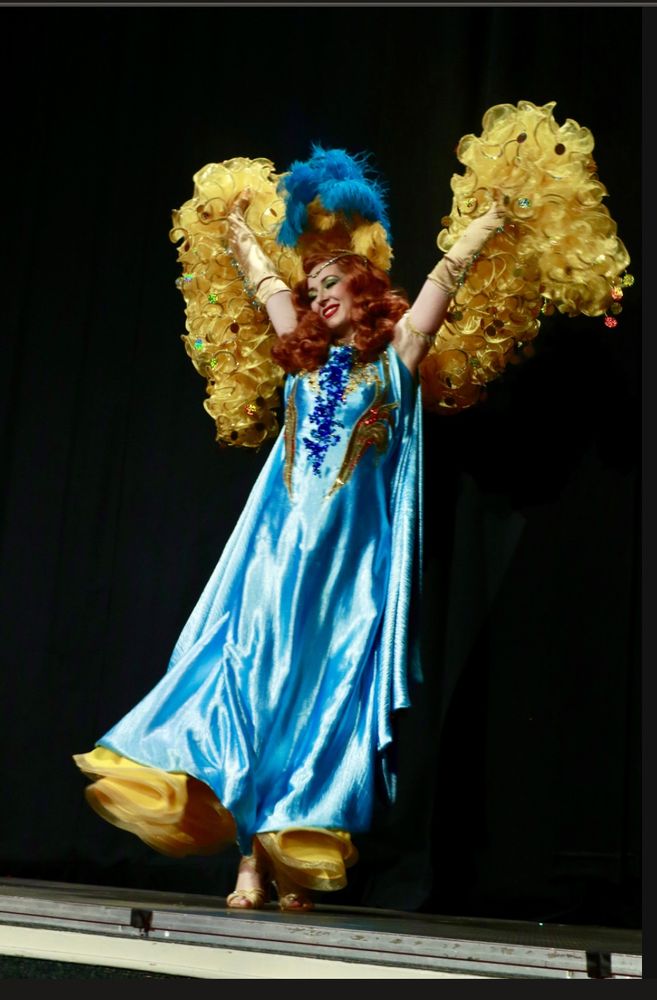 A fabulous burlesque performer on stage with a long light-blue gown, a feather headdress and a luscious yellow organza boa 