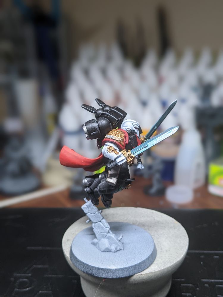 A raven guard jump captain leaping forward doff a rock.  He's wielding dual power swords.  His armor is deep black with gold details.  A flowing red cape trails behind him. 