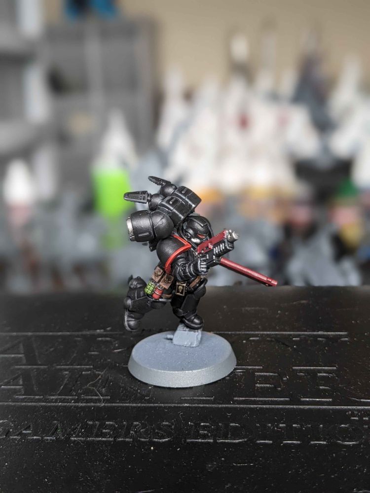 A painted Warhammer 40k miniature of a black-armored Space Marine with a jump pack, posed mid-leap. The model is holding a red power sword in its right hand and has various pouches and grenades on its belt. The base is unpainted gray, and the background is blurred, showing shelves and hobby supplies.