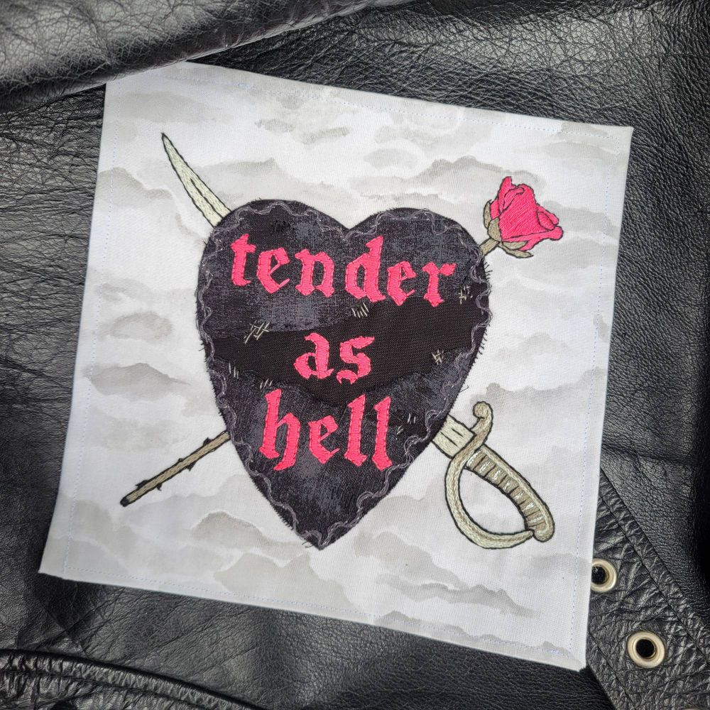 Photo of embroidered art featuring a black heart with the words "tender as hell" in hot pink and a painted background of gray clouds. The artwork is displayed on top of a black leather jacket.