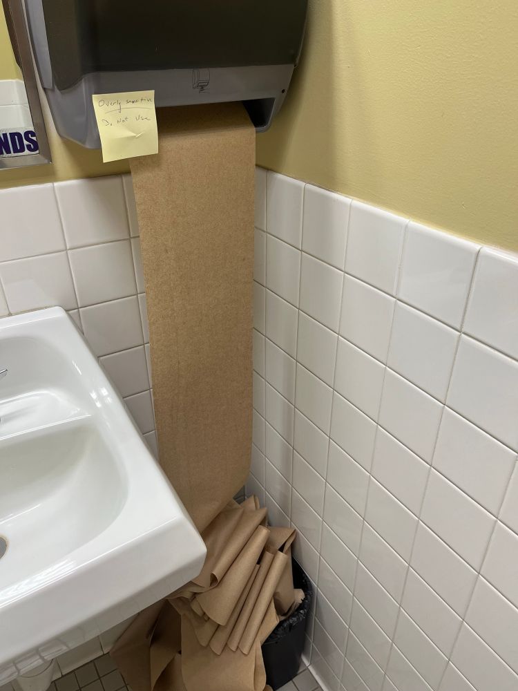 A photograph of a wall-mounted paper towel dispenser in a staff bathroom, with a Post-it note stuck to the machine that says “overly sensitive, do not use.” The machine has a single sheet of paper towel coming out of it approximately 10 feet long, puddled into a trashcan on the floor
