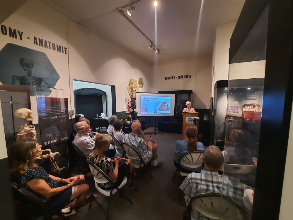 Woman at a lectern in front of about two dozen people, in a small room with medical displays including a skeleton. 