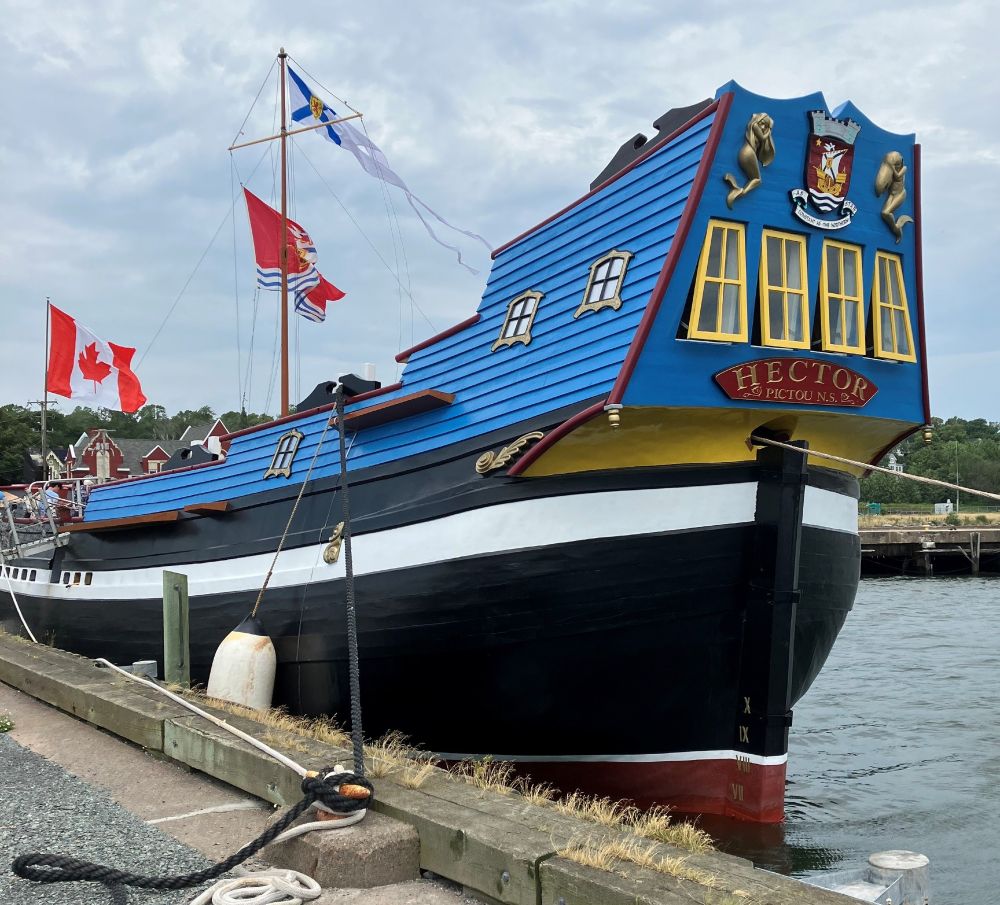 Photo of the back half of a tall ship replica, at a dock. The ship is relatively small, and painted bright blue above the deck. 