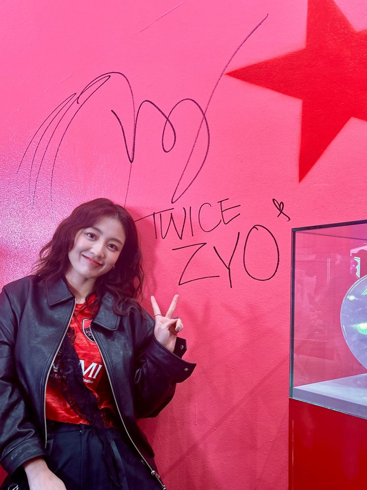 twice jihyo standing in front of a red wall with her signature that reads “twice zyo.” she is smiling a soft smile and holding up a piece sign