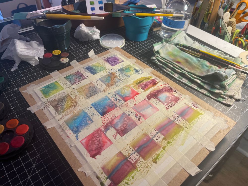 Square shaped small watercolor paintings with varying colors and splotchy shapes, masking tape blocks off the square painted areas on the paper