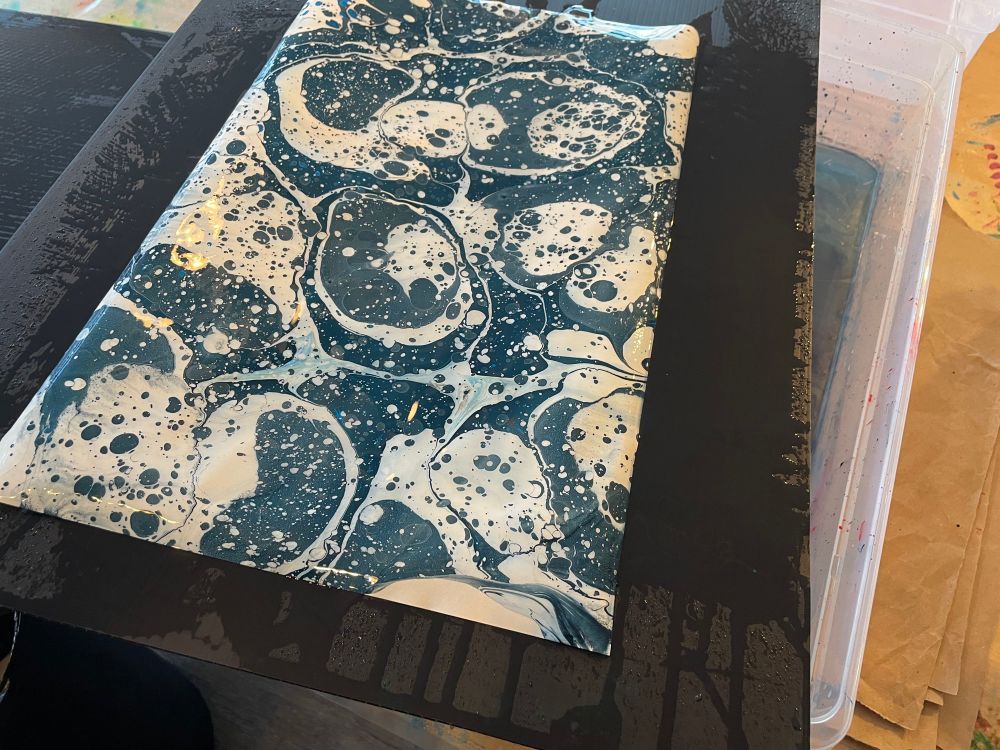 A “printed” sheet of marbled paper lies wet on a transfer board just after receiving the speckled and splotchy patterns of paint from the tray of size.