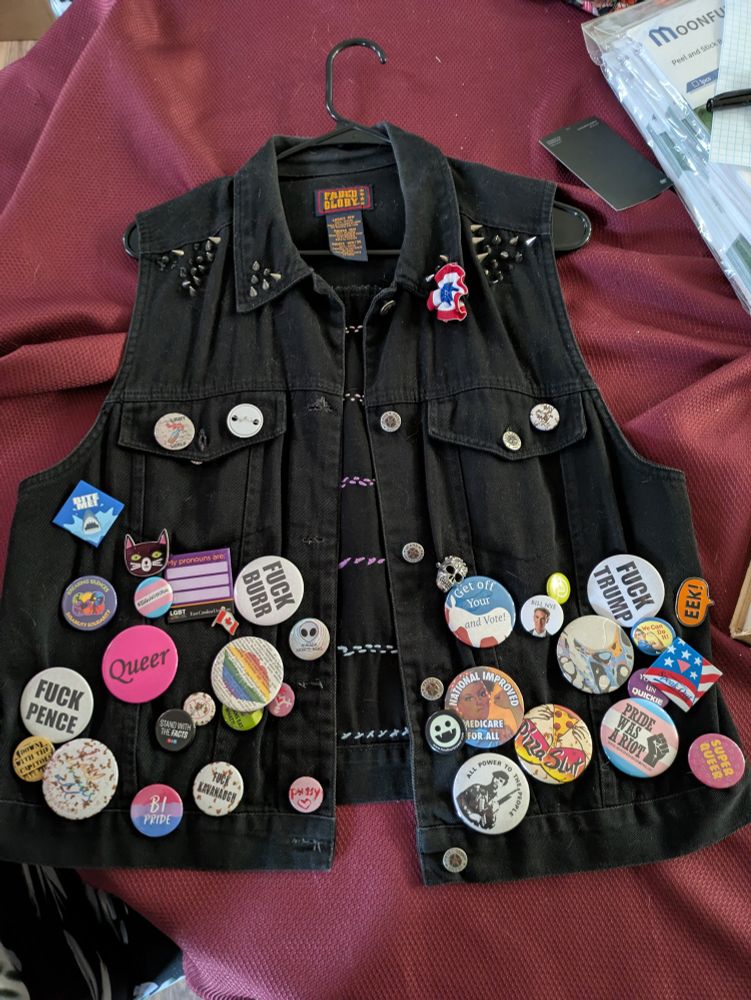 A black denim vest with spikes on the collar and dozens of pins, mostly leftist politics and queer support messages.