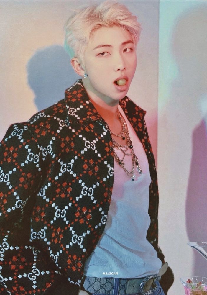 Namjoon with blond hair, wearing a patterned jacket over a white tank top with lots of jewelry, a single green grape between his luscious lips