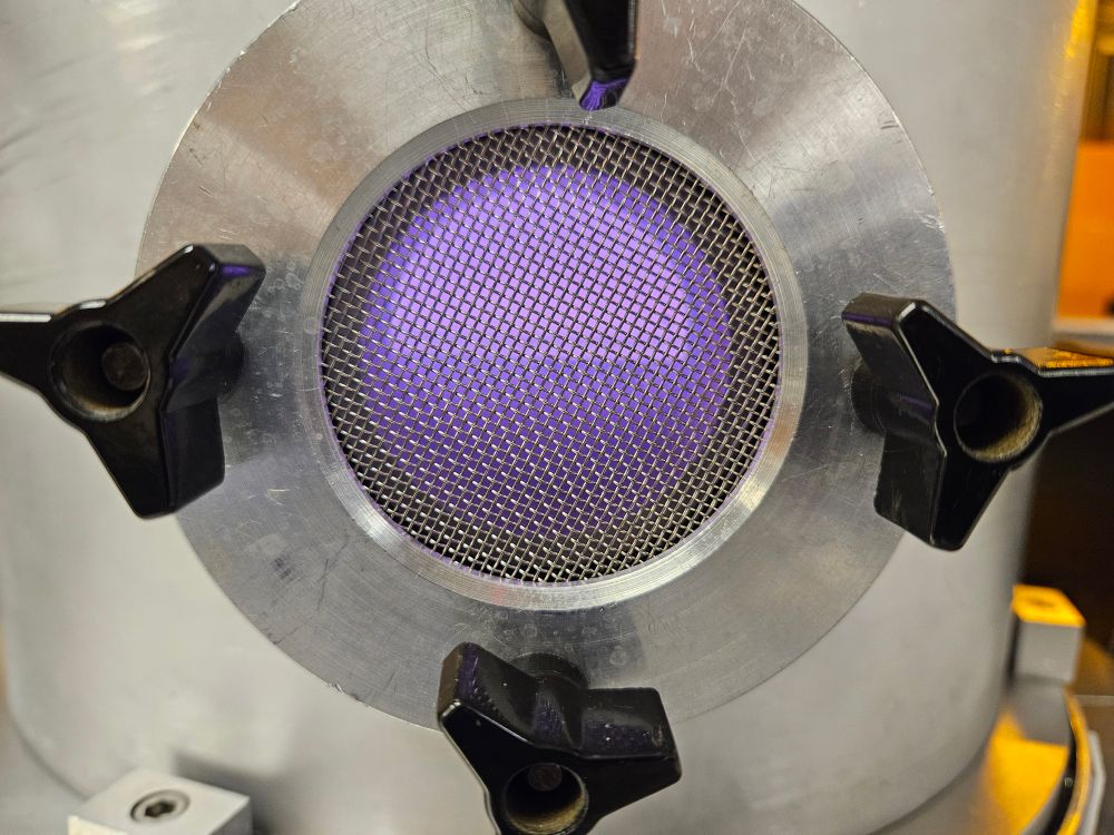 A metal chamber with a mesh covered window and a purple glow inside