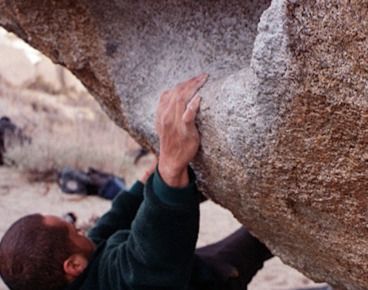 A man bouldering outdoors with his right hand on a chalked up sloper hold. 