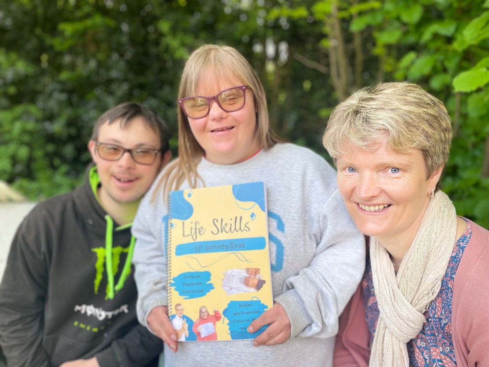 Adult lady with Down Syndrome holding a puzzle book saying Life Skills. Man with Down Syndrome and female teacher also in the picture.