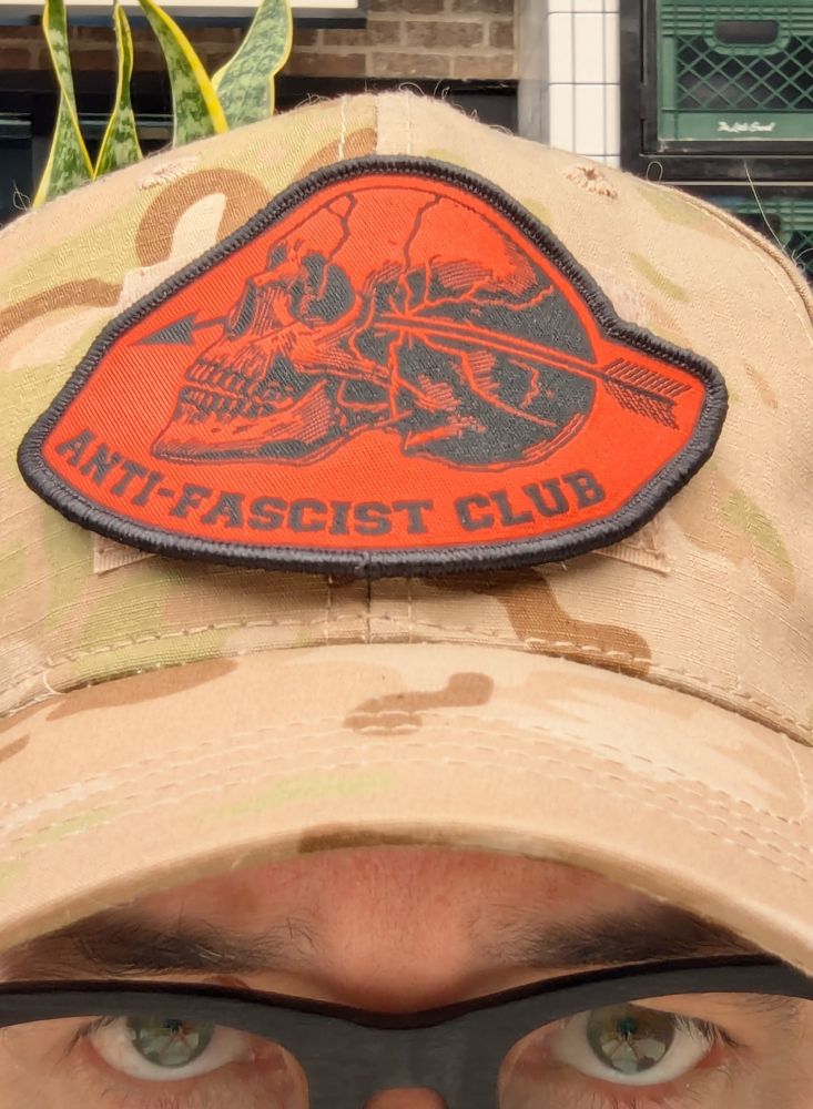 Photo of Brian in a camp hat with a red and black patch featuring a skull with an arrow through it reading "Anti-fascist Club"