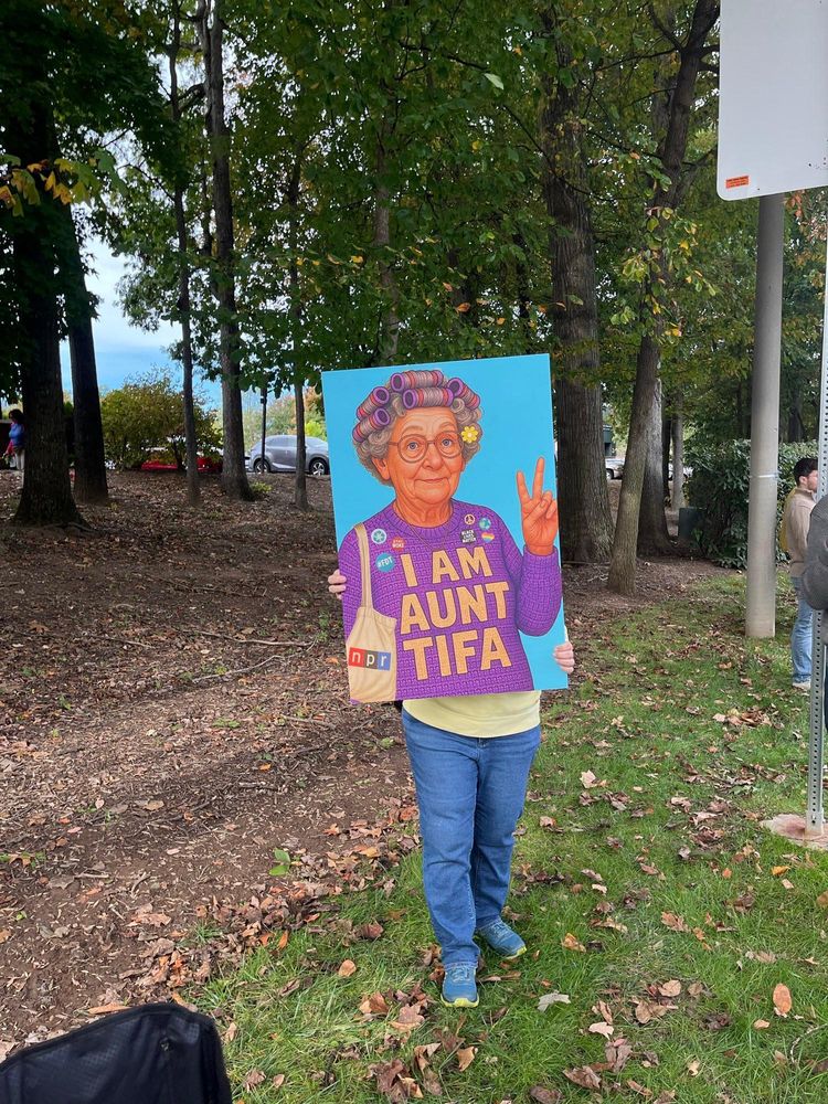A protester holding a sign that covers her upper body in a way that looks like the image on the sign is her upper body. The sign shows the upper body of an older woman in hair curlers. She is making a peace sign with one hand, and her sweater reads, "I am Aunt Tifa."