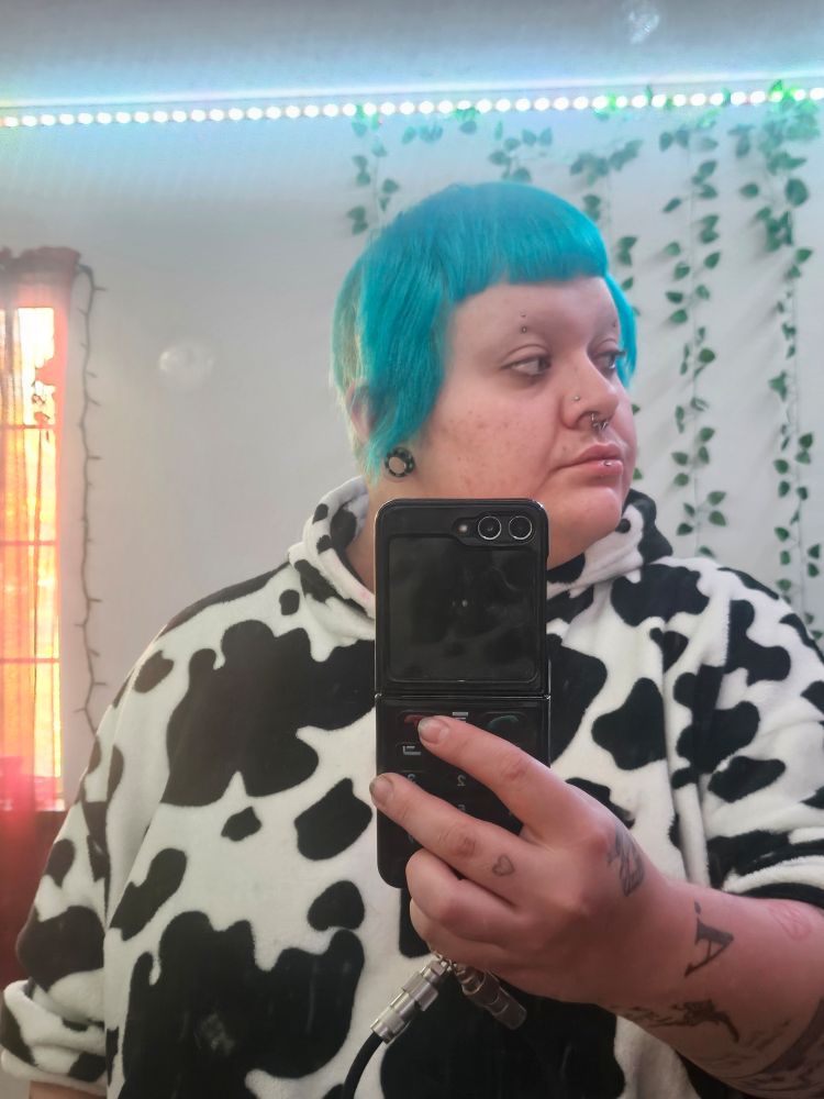 A mirror selfie of clyde wearing an oversized fleece hoodie that has cow print on it. His hair is cut into a Chelsea cut-- shaved in the back and long bangs framing the face. His hair is alls dyed bright blue.