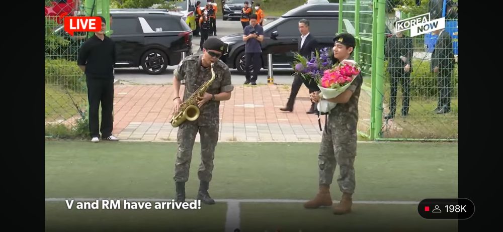 Namjoon plays saxophone while V looks on smiling 