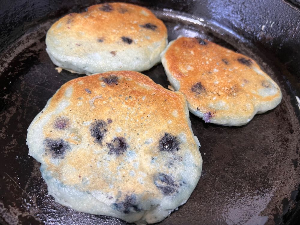 Cast iron skillet with three golden brown blueberry pancakes (or, flat blueberry muffins, if you like!)