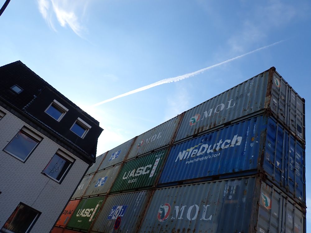 Unter blauem Himmel rechts eine Wand aus verschiedenfarbigen Containern, links ein schräg nach rechts geneigtes Wohnhaus.