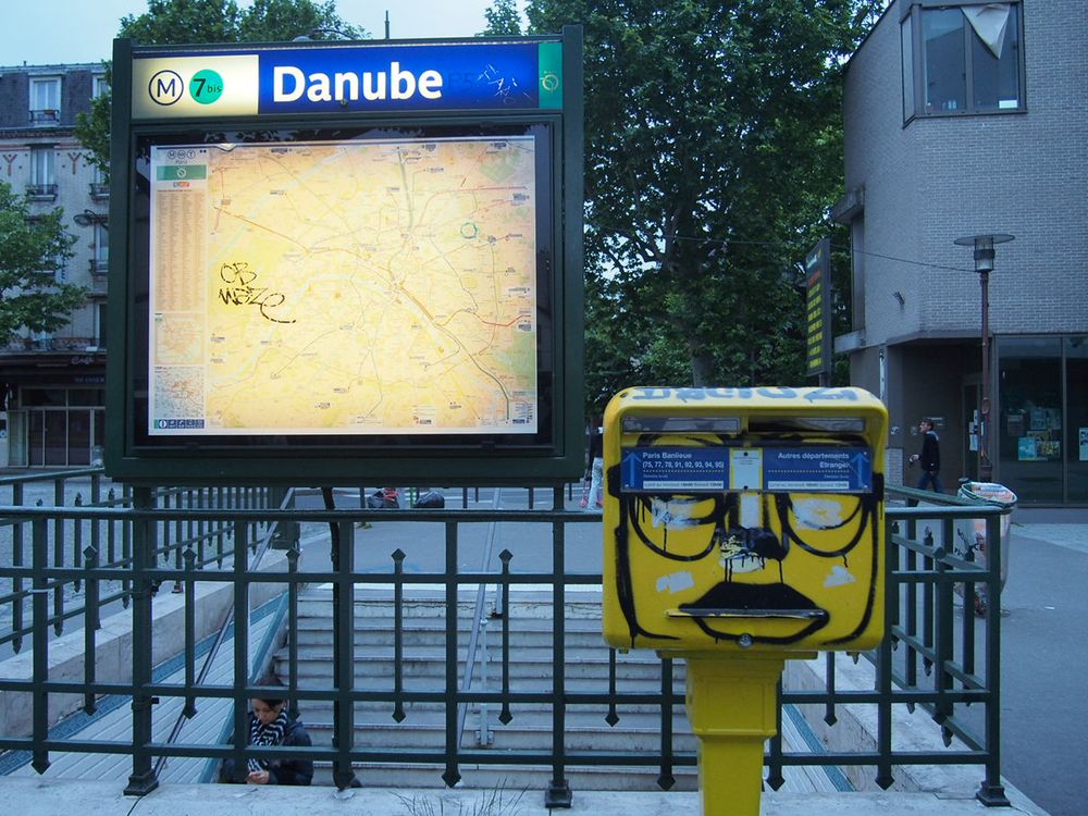 Urbane Szene. Links Schaukasten mit Stadtplan, erleuchtet, oben in weiß auf Blau Hinweis auf Metrostation "M7bis Danube". Recht daneben ein gelber Briefkasten auf gelber Säule, mit Graffitti verziert, ein Gesicht darstellend. Dahinter ein Metallgeländer und Treppenabgang zur Metro.