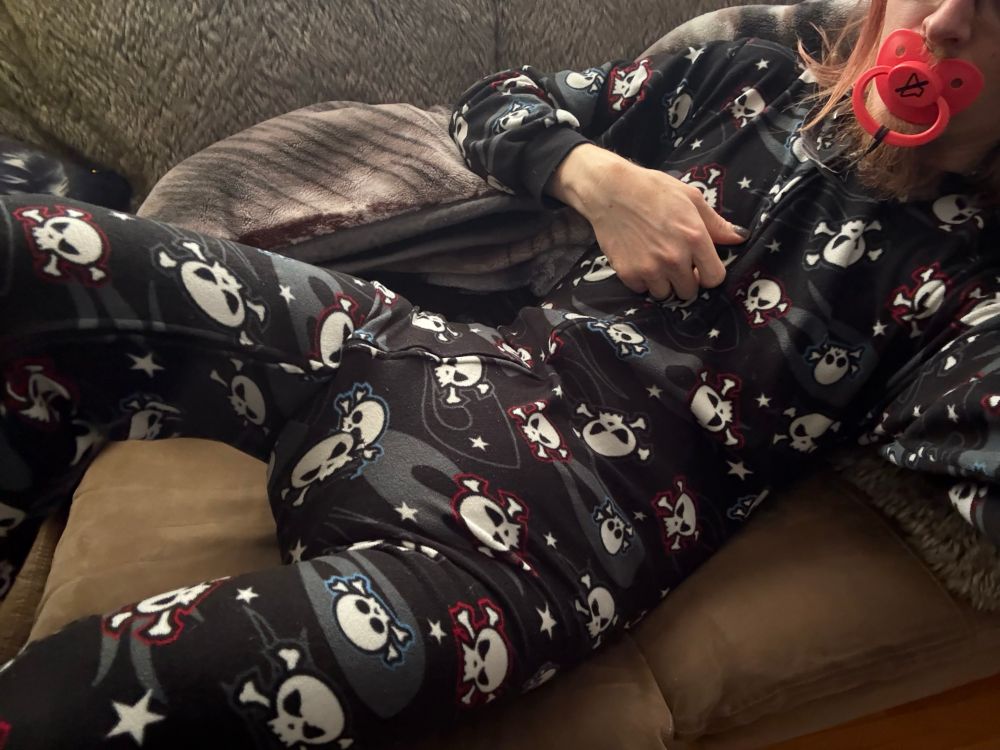 A little ABDL diaper pup laying in the couch on a Saturday morning wearing footie pajamas with skulls and bones all over them against a black background with a bright red mute button pacifier planted in his muzzle. 

The footies have a zipper that goes from the ankle to the neck, and barely contain the tell-all bulge of a plump “pumpkin-butt” diaper. 
