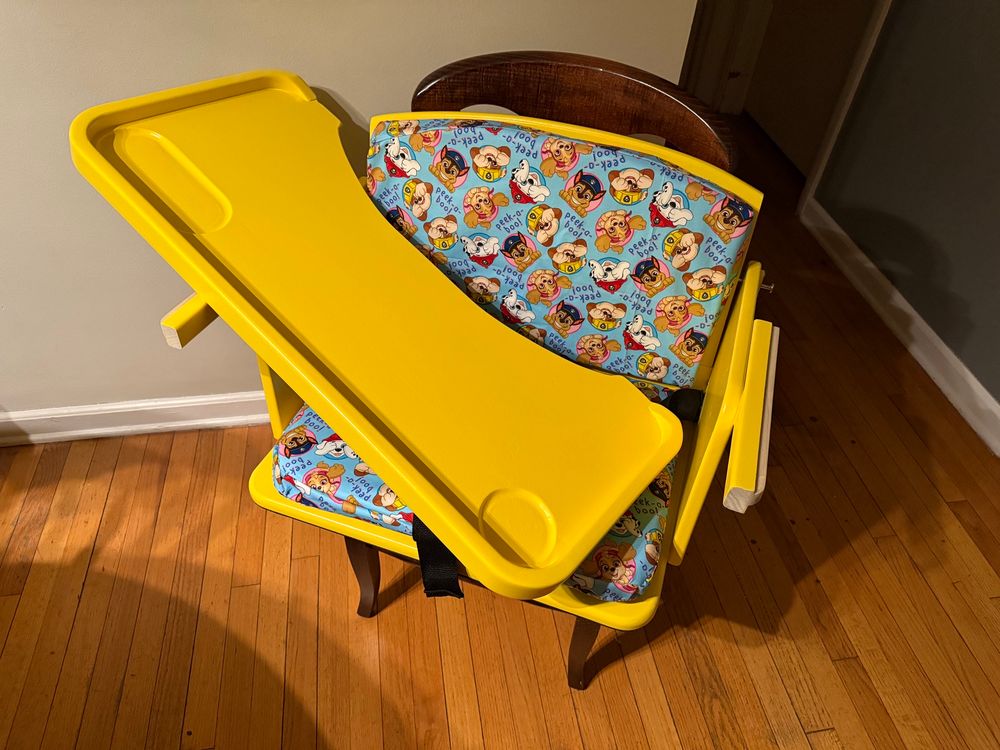 A bright yellow wooden seat with paw patrol themed cushions in blue. The chair is strapped to a regular dining room chair and has a matching removable tray with a raised lip around the edge and hollowed out spaces for snacks or a drink. The tray is sitting inside the seat, off its tracks at the moment. 