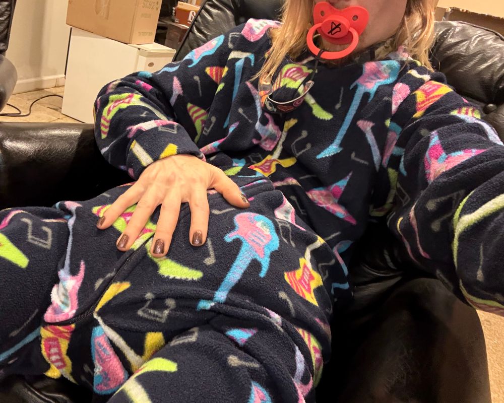 A long haired femboy with purple painted nails wearing a fleece footie pajama with a zipper all the way down the leg further making a resemblance to a toddler footie pajama set ,covered in electric guitars and music notes, he’s got a bright red mute button pacifier in his muzzle connected to his footies by a spooky pacifier clip. 

Barely contained by the footies is the obvious bulge of a huge diaper with the “painted claws and paw” of the femboy diaperpup resting on the bulging diaper beneath the confines of his footie pajamas.