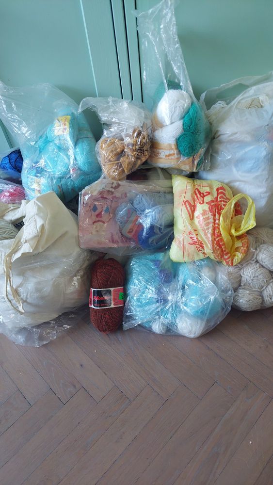 A big pile various plastic bags filled with skeins of wool and cotton in various light colours.