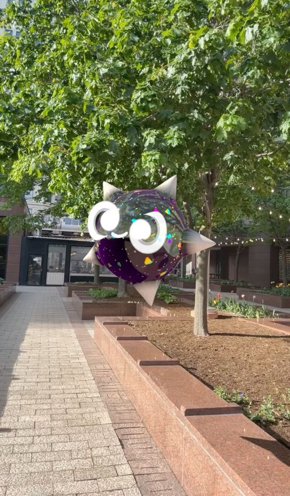 A 3D render of the shiny Pokémon Minior (core form) in the real world outdoors. Minior is a glitter-filled ball shape with 5 protruding points like a star, two white swirly eyes and a smile. It hovers above a brick walkway by a leafy tree.