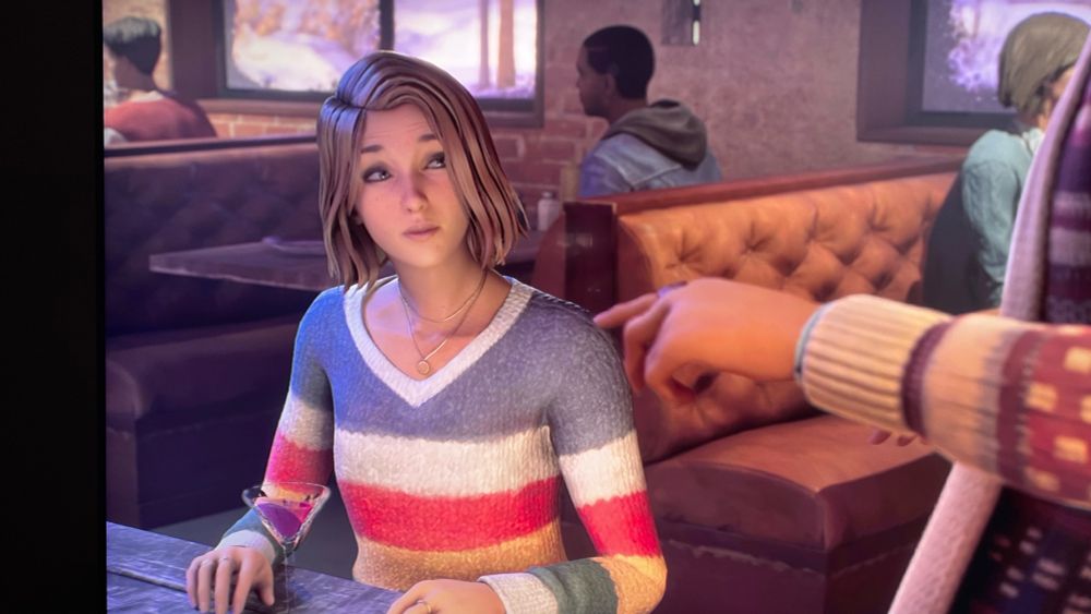 Screenshot of Life is Strange game. Max in a striped rainbow sweater, sitting in a cafe. Early part of the game.