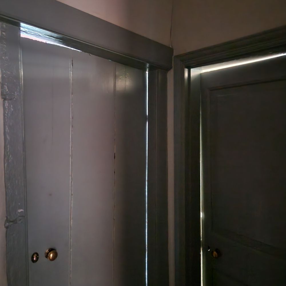 Two grey doors with brass handles in a dark corridor. There's clear triangles of light around the tops of both frames, because neither frame is even slightly square 