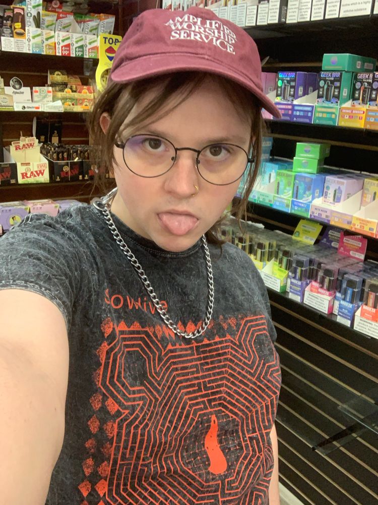 a woman posing with her tongue out in front of a wall of vapes. she is wearing a steel chain, black rimmed glasses, a red hat with white letters that reads, “AMPLIFIER WORSHIP SERVICE,” and a gray shirt with red writing that reads “slowdive”