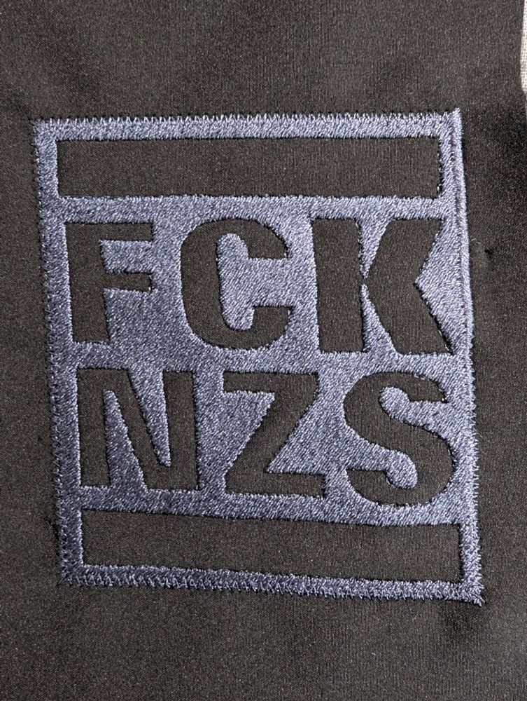 Das Bild zeigt einen rechteckigen, aufgenähten Aufnäher auf einem dunklen, möglicherweise schwarzen Stoff. Der Aufnäher ist rechteckig und hat einen grauen Rand, der aussieht, als wäre er gestickt. Innerhalb des Rechtecks sind die Buchstaben "FCK" in einer Zeile und "NZS" in der Zeile darunter zu sehen. Die Buchstaben sind ebenfalls grau und scheinen gestickt zu sein. Zwischen den Buchstaben und dem Rand des Aufnähers befinden sich graue, horizontale Balken. Der Hintergrund des Aufnähers ist ebenfalls grau. Das gesamte Design wirkt einfach und kontrastiert mit dem dunklen Stoff, auf dem es angebracht ist.